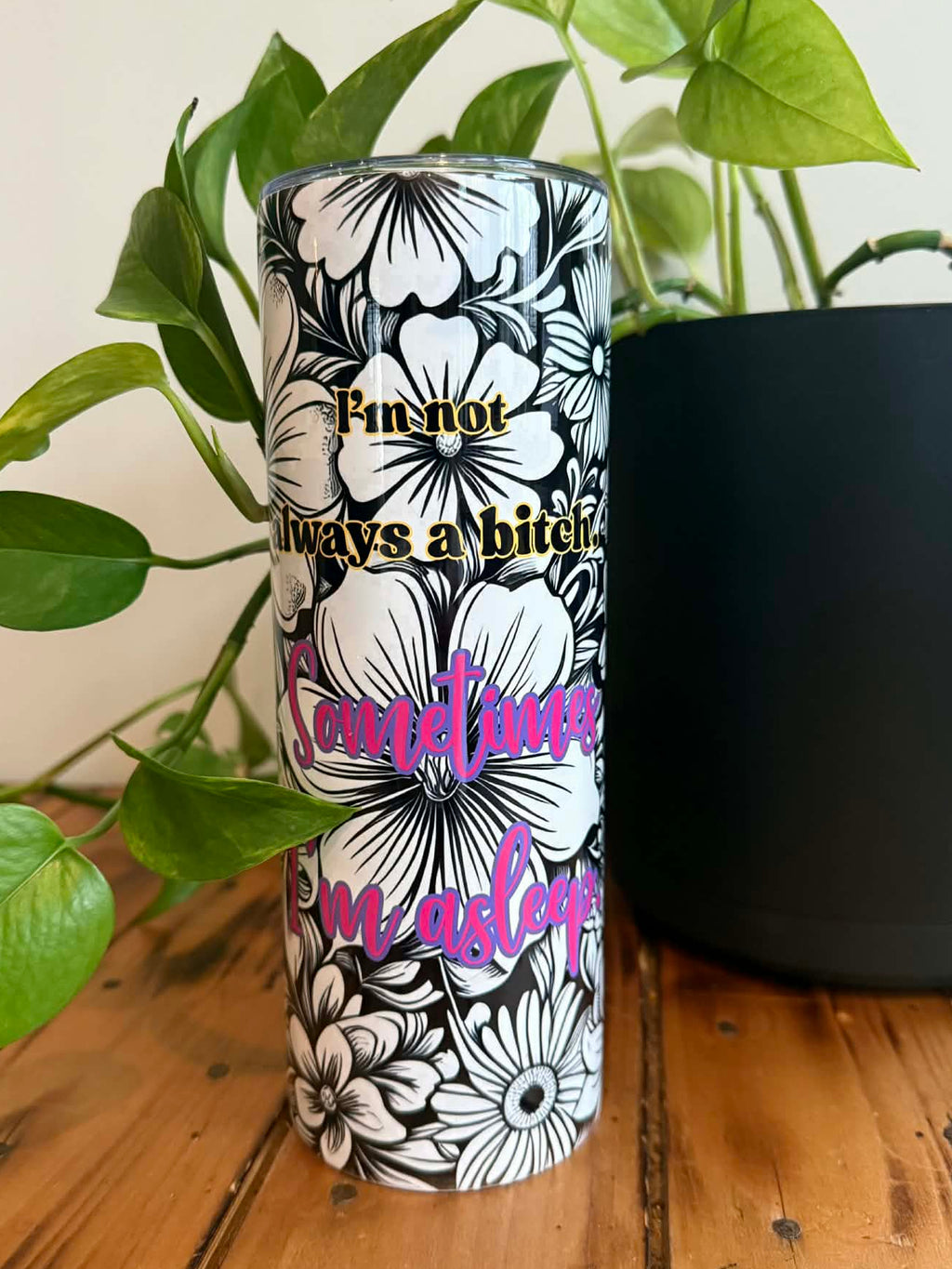 Floral-patterned tumbler with text on a wooden surface with a plant in the background