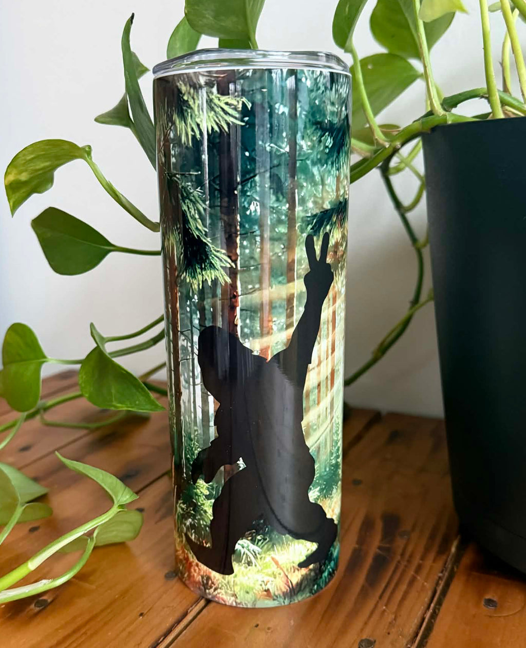 Tumbler with sasquatch silhouette design in the woods on a wooden surface with plants in the background