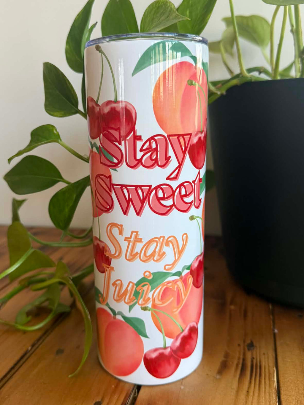 Tumbler with 'Stay Sweet, Stay Juicy' text and fruit graphics on a wooden surface with plants in the background