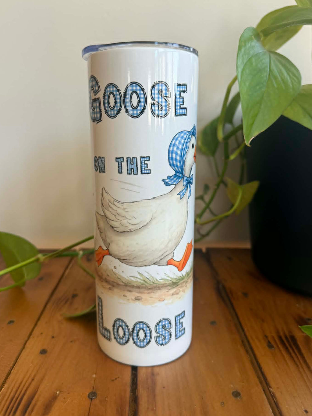 Tumbler with a goose design and text on a wooden surface with plants in the background