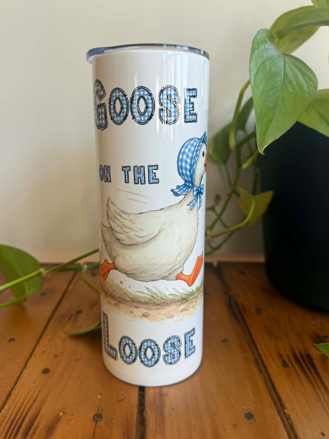 Tumbler with a goose design and text on a wooden surface with plants in the background