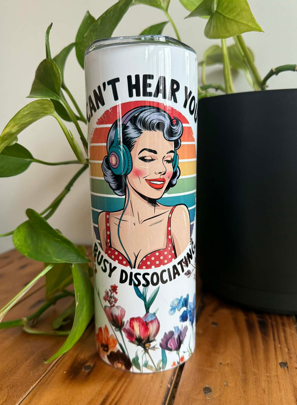 Tumbler with a retro style design of a woman wearing headphones and text with flowers, placed on a wooden surface with plants in the background.