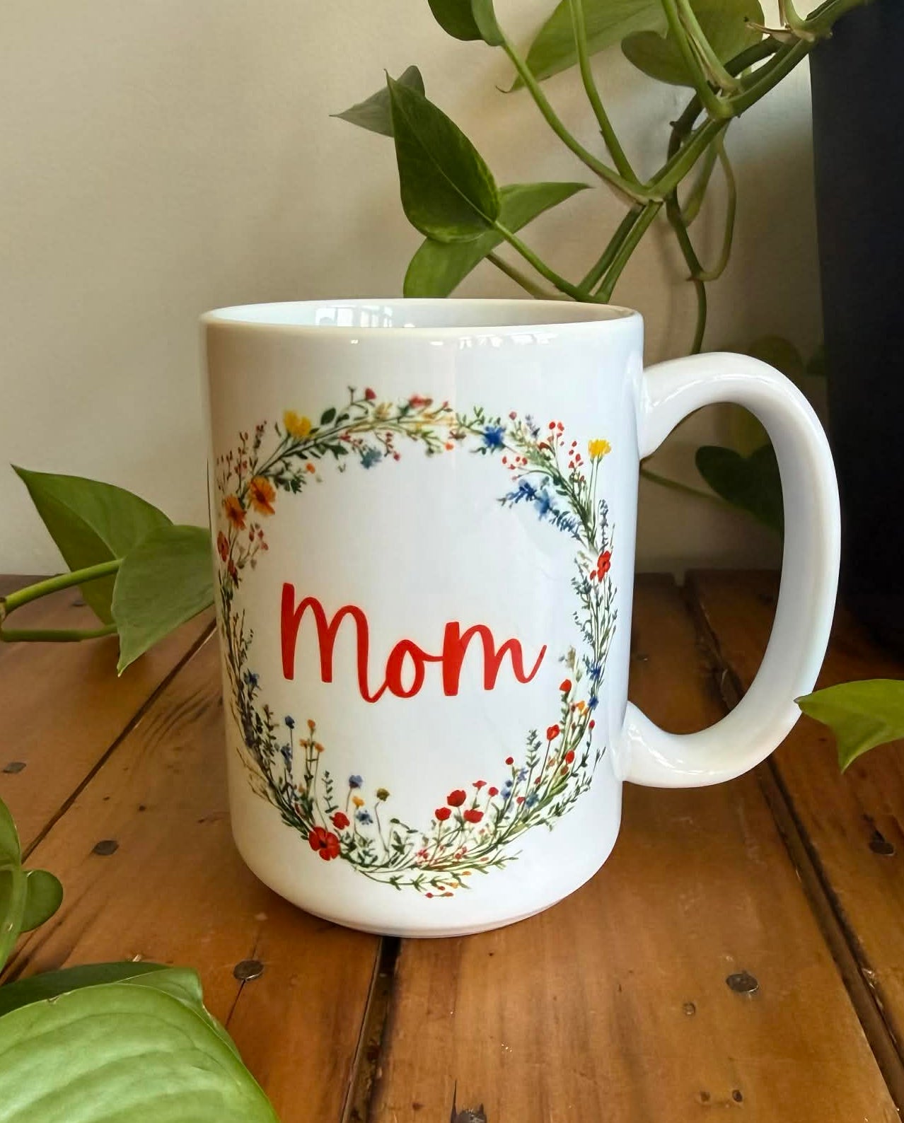 White mug with floral design and 'Mom' text on a wooden table with plants.