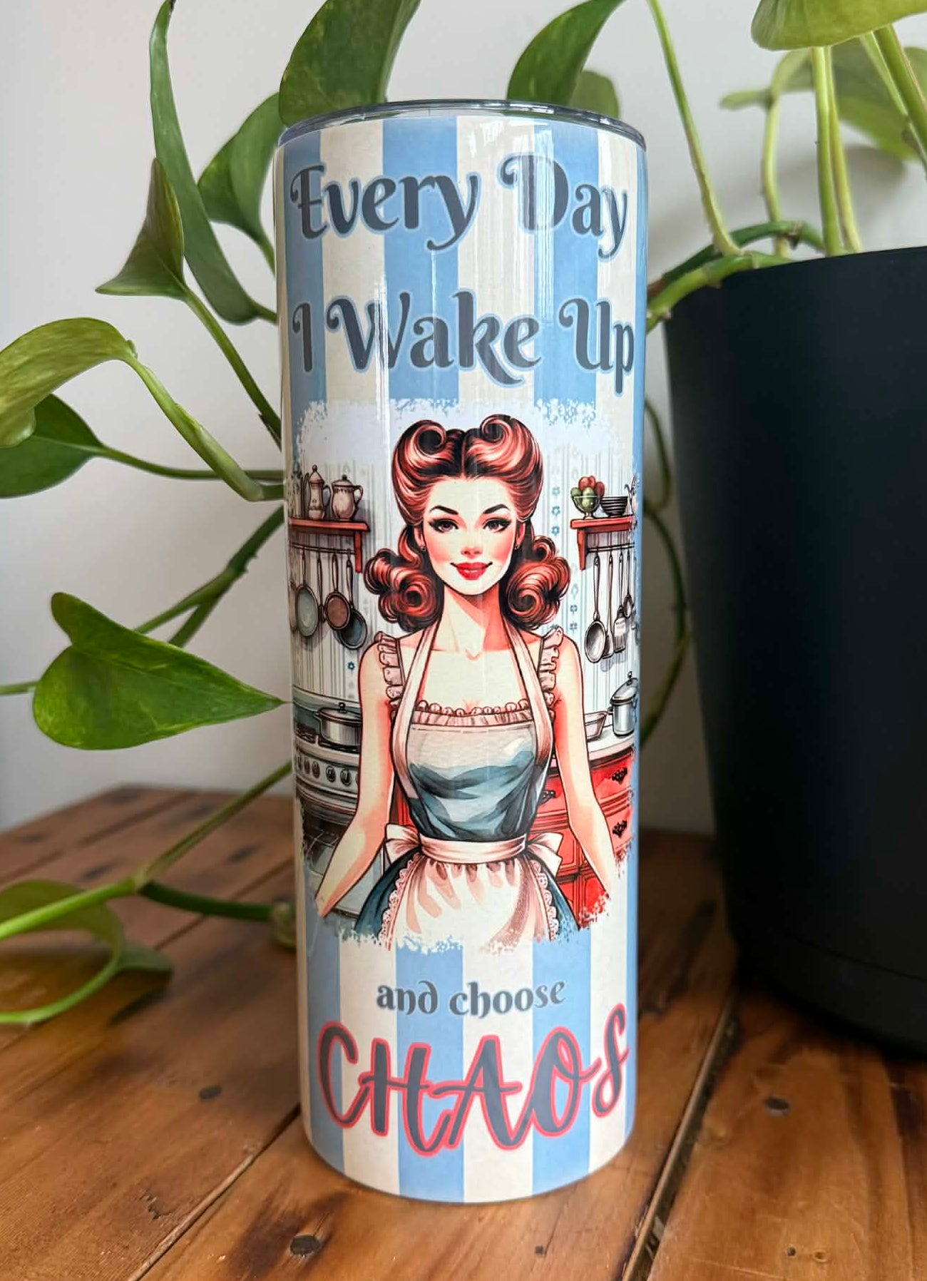 Tumbler with a retro design of a woman in a retro kitchen and text 'Every Day I Wake Up and Choose Chaos' on a wooden surface with plants.