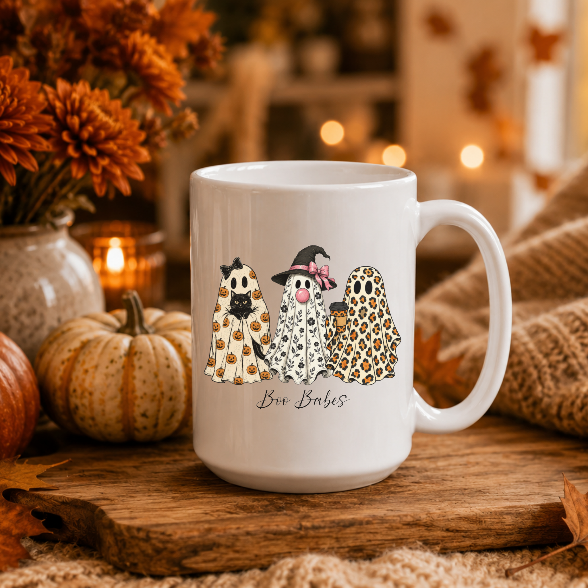 A white ceramic mug with a Halloween-themed graphic of three ghosts in costumes, placed on a wooden surface with autumn props like pumpkins, dried flowers, and a cozy blanket in the background.