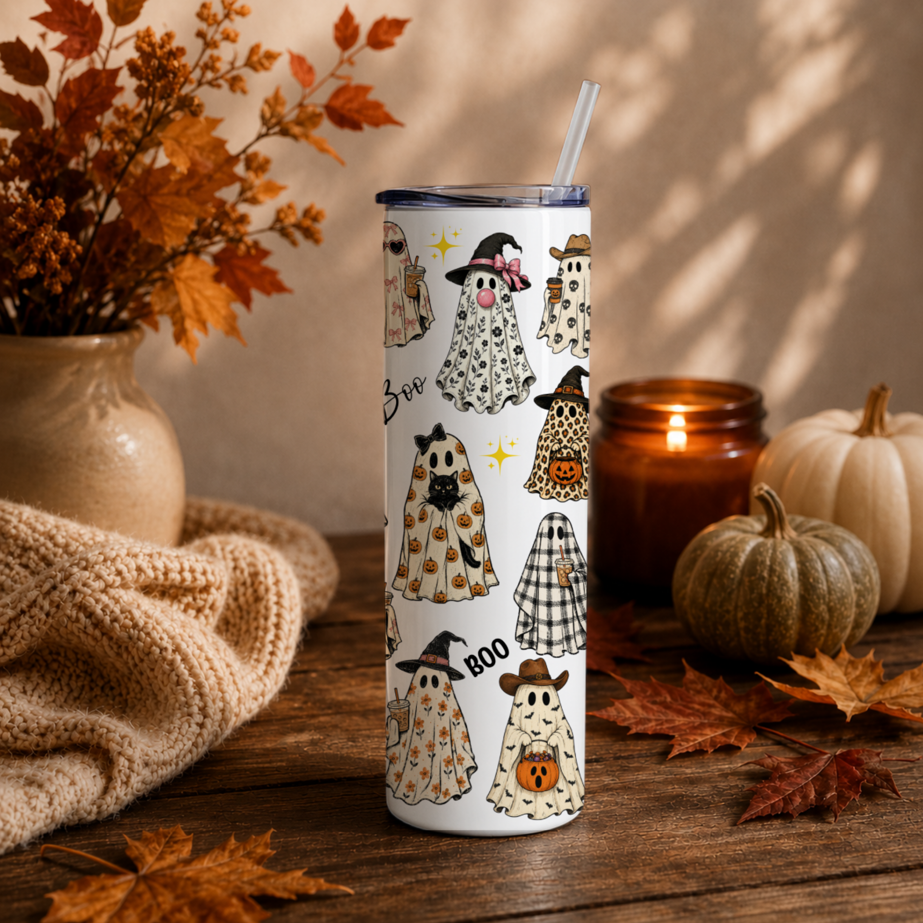 A tall white 20 oz insulated tumbler with a whimsical Halloween ghost pattern, a clear lid, and a clear straw, set on a rustic wooden table surrounded by autumn leaves, a knitted blanket, pumpkins, and a lit candle.