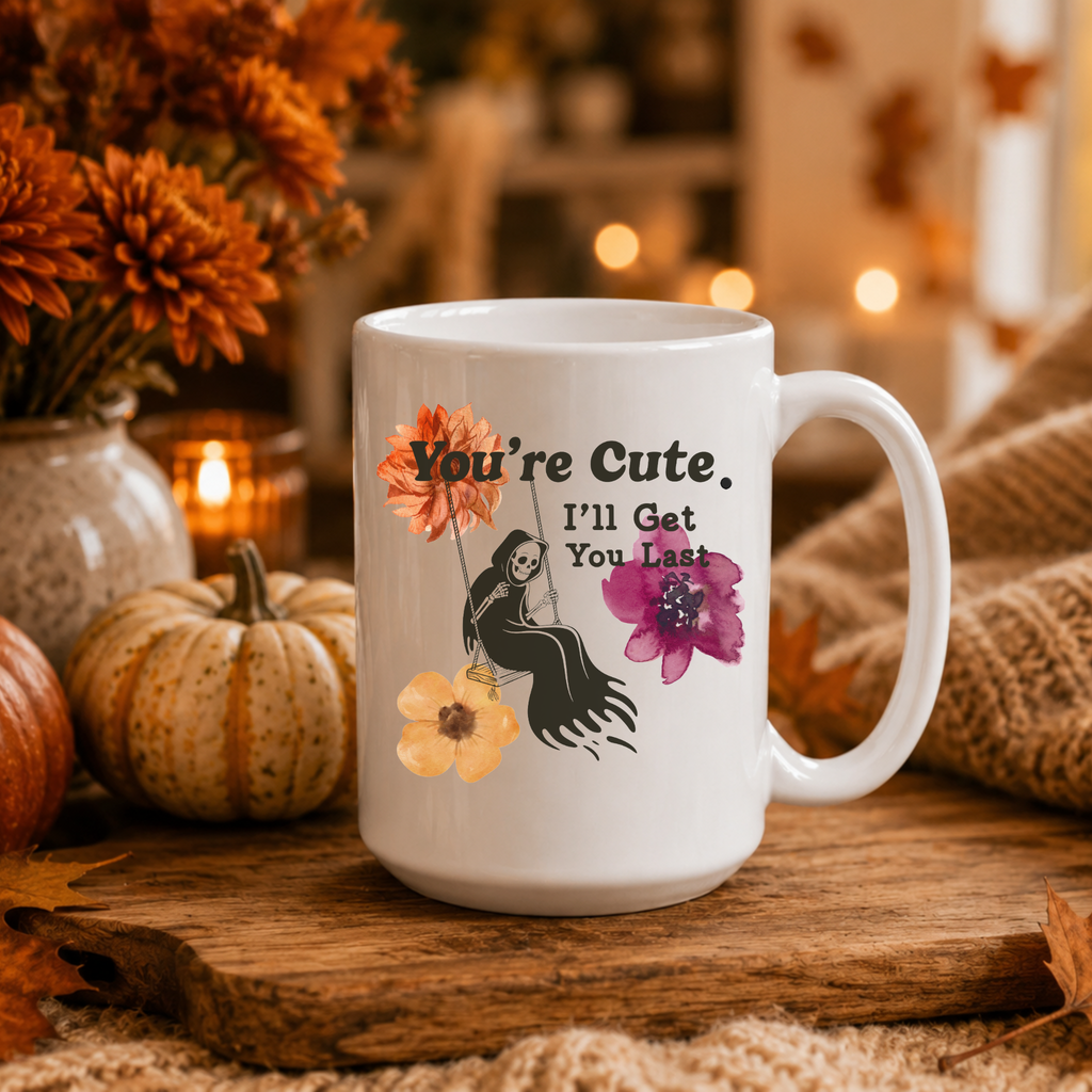 A white ceramic coffee mug printed with the text 'You're Cute. I'll Get You Last' and an illustration of a grim reaper on a swing with orange, yellow, and purple flowers.