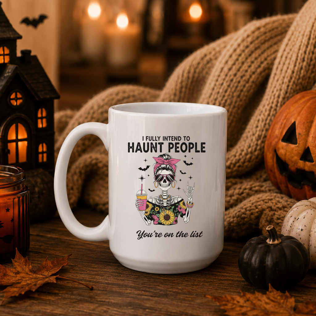 A white ceramic mug with a sassy and fun skeleton holding a coffee cup and a peace sign surrounded by bats with the text "I fully intend to haunt people - You're on the list" placed on a wooden surface surrounded by Halloween decorations
