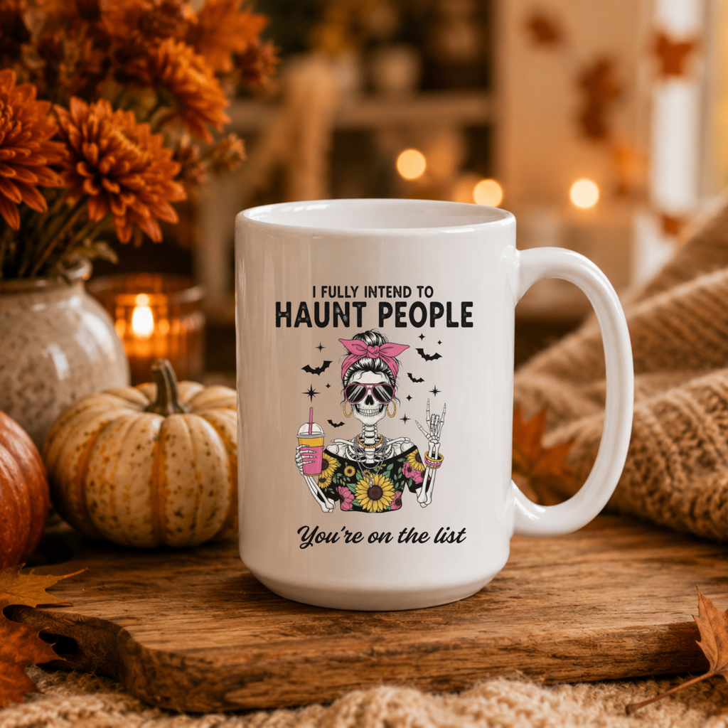 A white ceramic mug with a Halloween graphic of a skeleton in a pink bow and a sunflower top, surrounded by bats and the text 'I FULLY INTEND TO HAUNT PEOPLE You're on the list', set on a wooden surface with autumn decorations including pumpkins, flowers, and candles in the background.