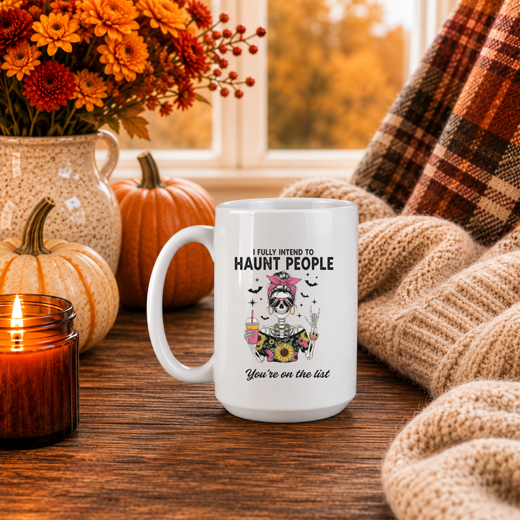 A white ceramic mug with a Halloween-themed graphic sits on a wooden table in a cozy autumn setting. The mug features a skeleton wearing a pink bow and holding a coffee, with the text 'I FULLY INTEND TO HAUNT PEOPLE' and 'You're on the list'.
