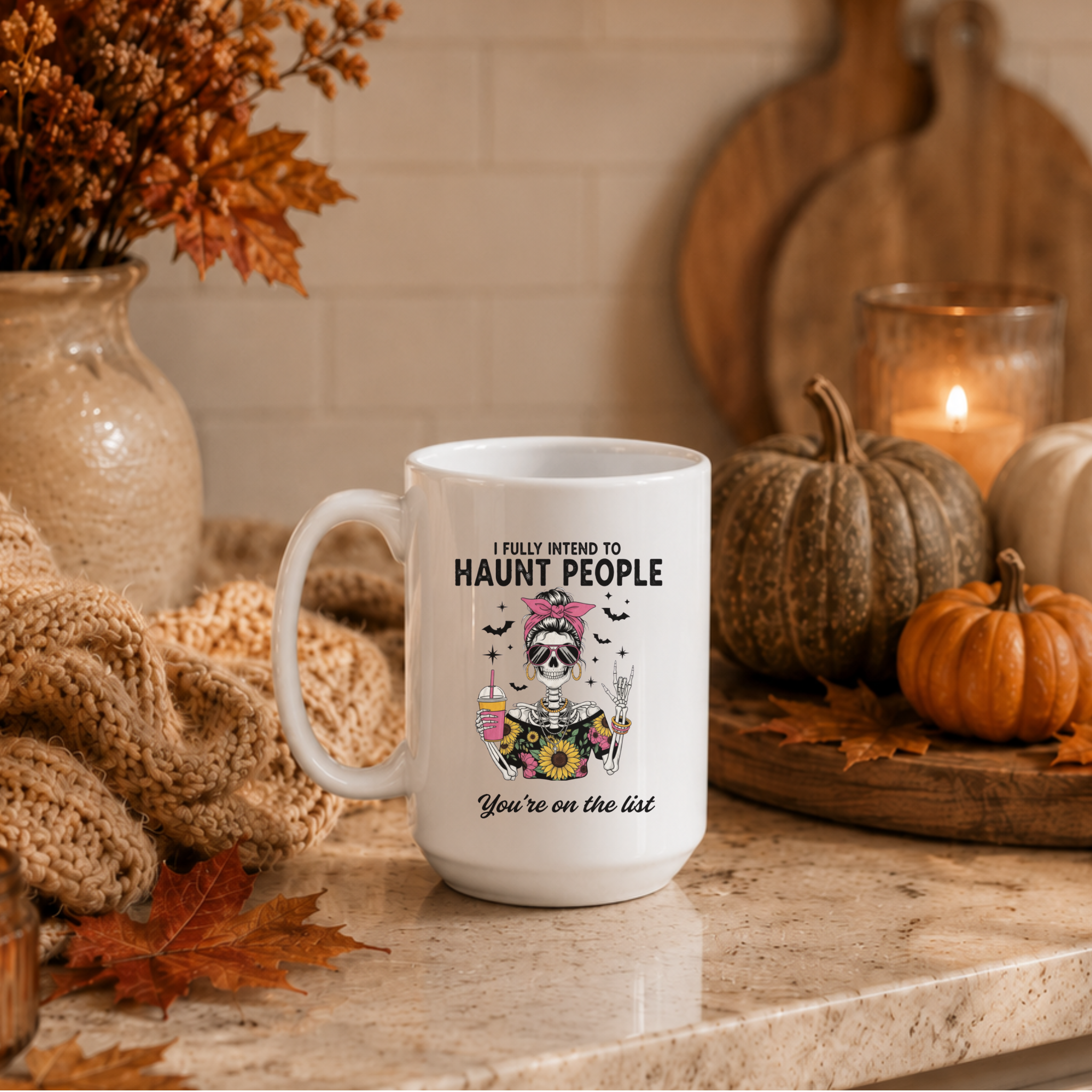 A white ceramic mug with a Halloween-themed graphic and the text 'I FULLY INTEND TO HAUNT PEOPLE You're on the list' sits on a kitchen counter surrounded by autumn decorations including pumpkins, a candle, and a knitted blanket.