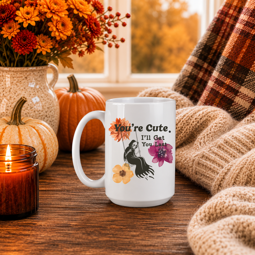 A white mug with a Spooky Image and Fun Flowers that say's "You're cute. I'll get you last" on a wooden table. 
