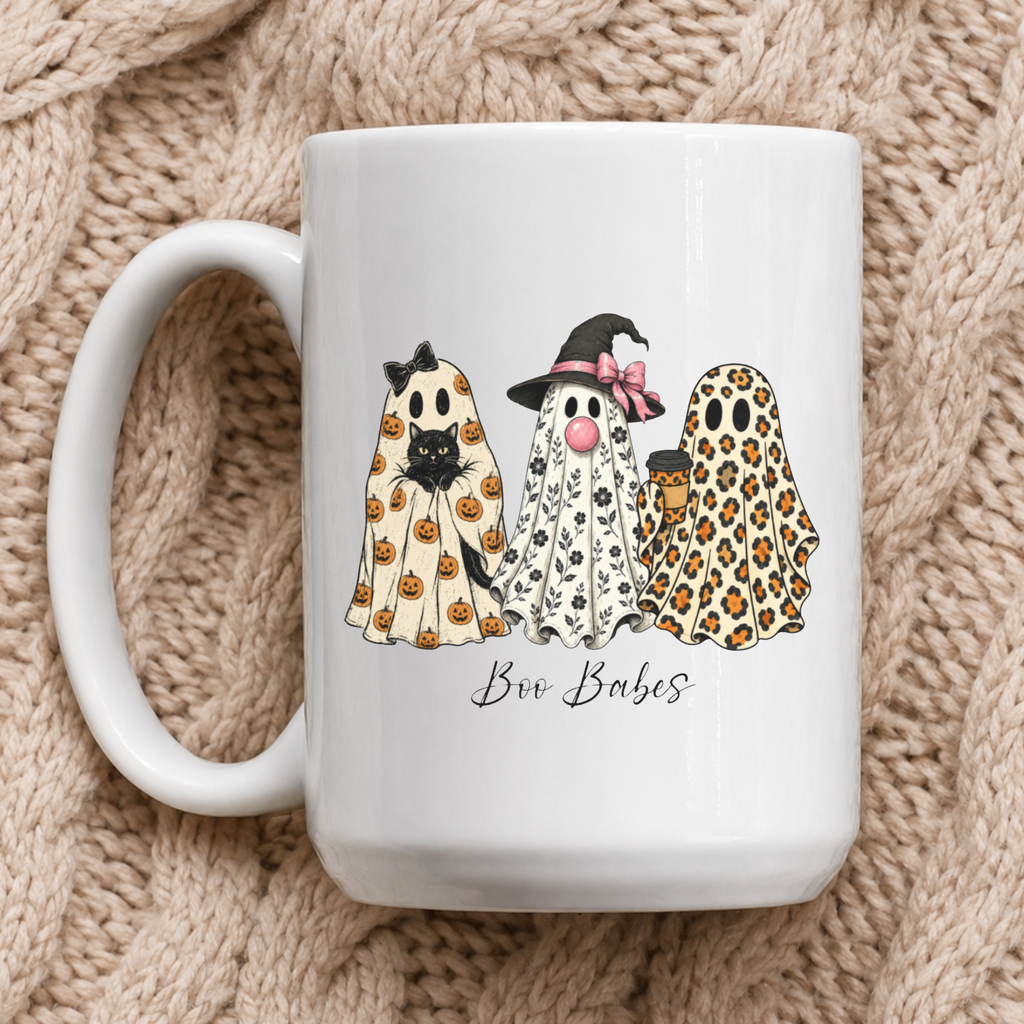 A white ceramic coffee mug featuring a cute illustration of three ghost friends in Halloween costumes (pumpkin print, black and white print, leopard print) with the text 'Boo Babes' underneath, set against a cozy beige knitted blanket background.