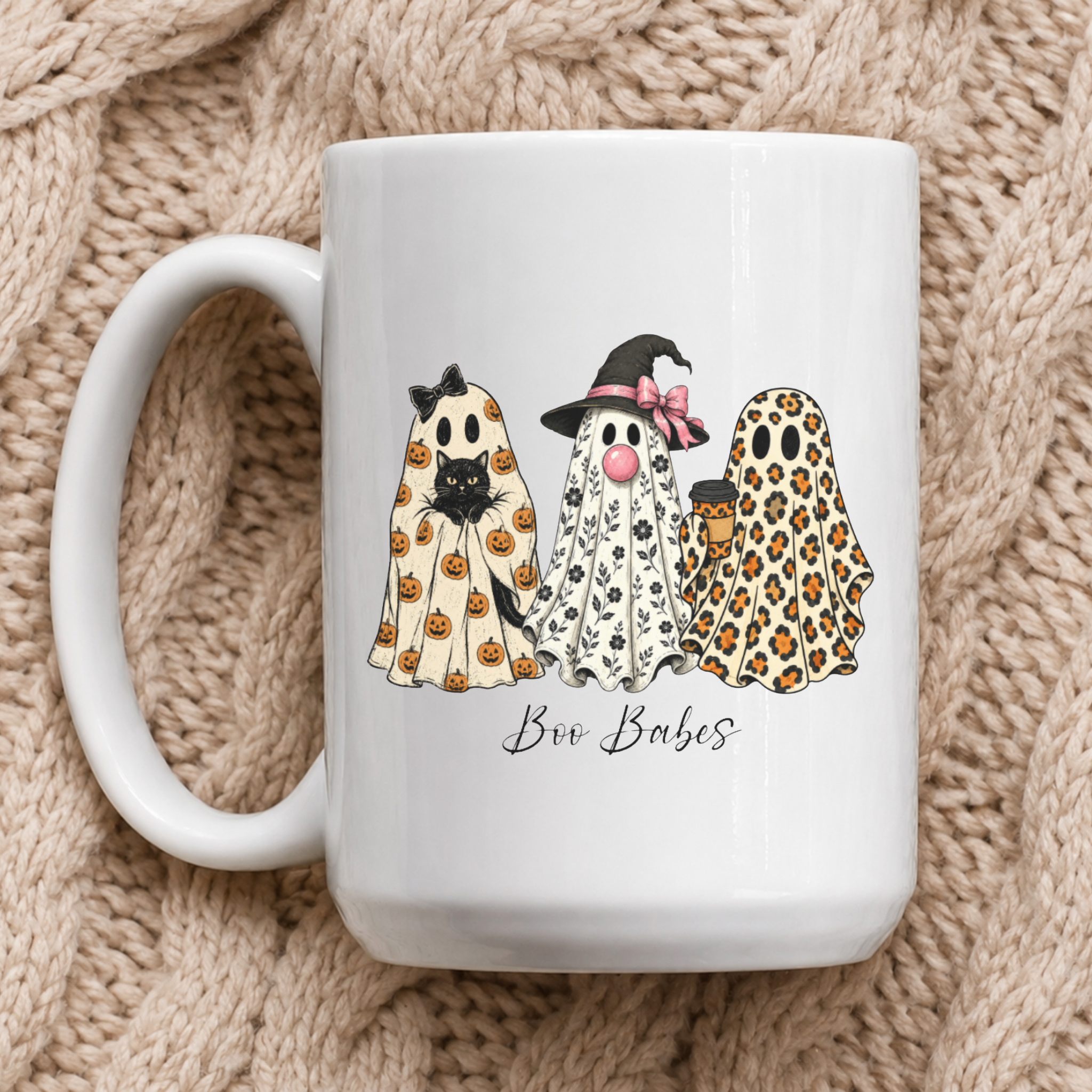 A white ceramic coffee mug featuring a cute illustration of three ghost friends in Halloween costumes (pumpkin print, black and white print, leopard print) with the text 'Boo Babes' underneath, set against a cozy beige knitted blanket background.
