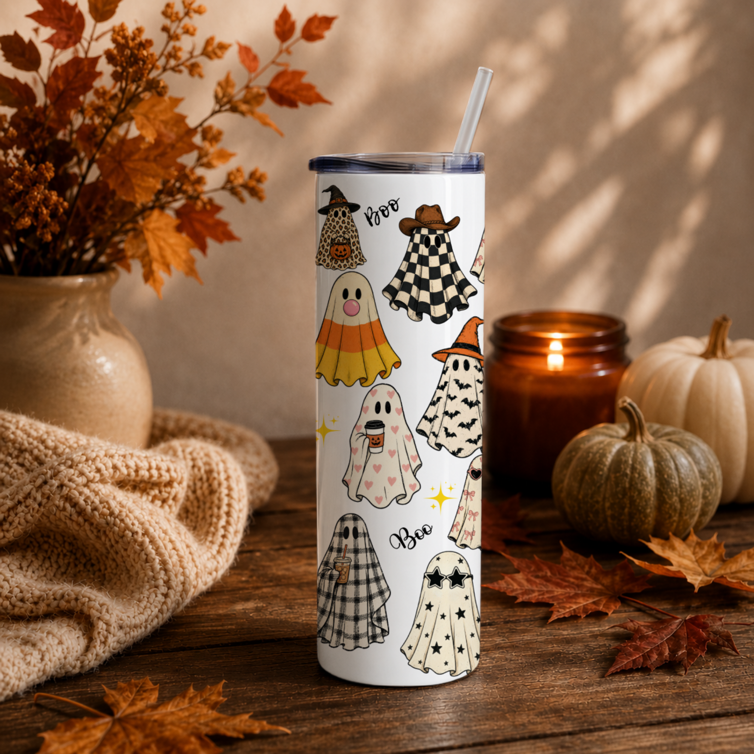 A white insulated 20 oz tumbler with a clear lid and straw, decorated with a Halloween pattern of ghosts, witches' hats, and plaid cloaks, styled on a wooden table with autumn leaves, a candle, and pumpkins.