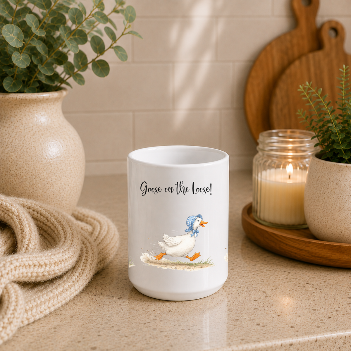 A white ceramic mug with a printed illustration of a goose and the text 'Goose on the Loose!' sits on a beige countertop in a styled kitchen setting with a candle, plants, and a woven throw nearby.