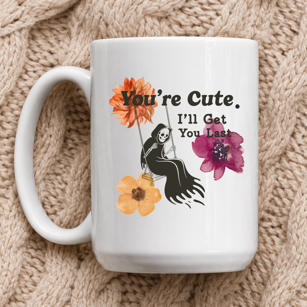 A white ceramic mug with a cute Halloween-style graphic, resting on a beige chunky knit blanket. The graphic features a black skeleton sitting on a swing with the text 'You're Cute. I'll Get You Last"
