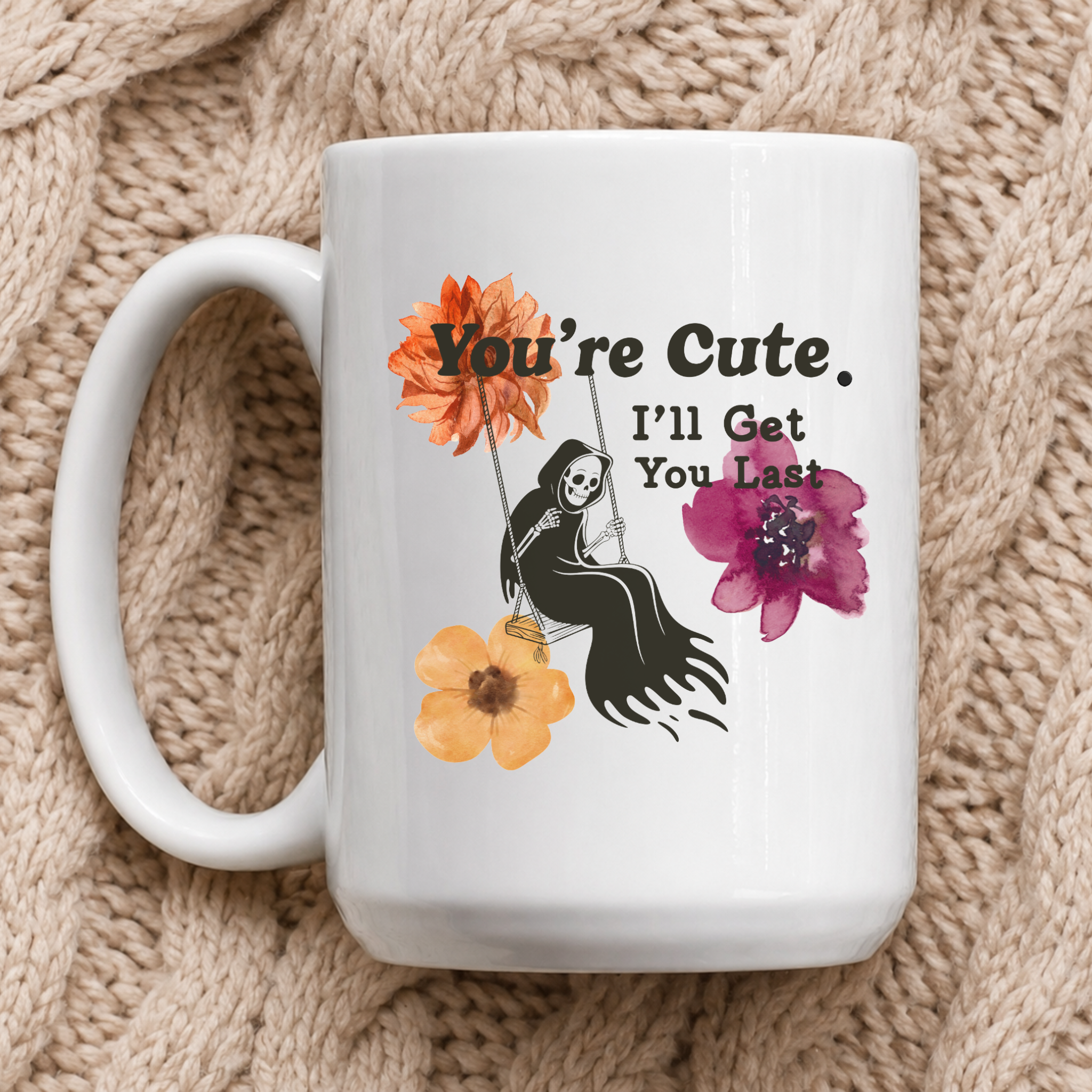 A white ceramic mug with a cute Halloween-style graphic, resting on a beige chunky knit blanket. The graphic features a black skeleton sitting on a swing with the text 'You're Cute. I'll Get You Last"