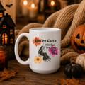A white ceramic mug with a Halloween-themed graphic print, styled on a wooden table with a cozy autumn background featuring a knitted blanket, a jack-o'-lantern, a small pumpkin, and a black lantern.
