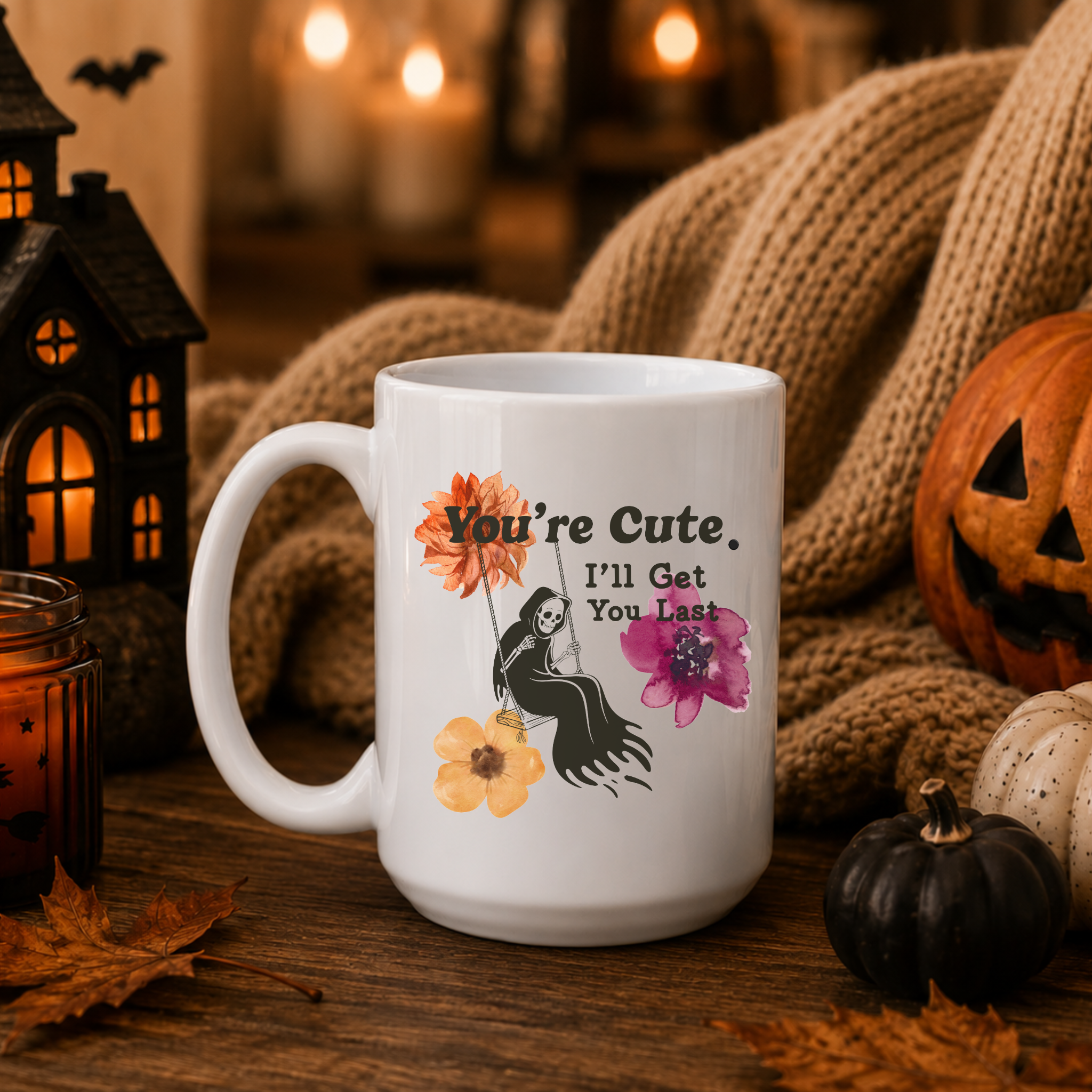 A white ceramic mug with a Halloween-themed graphic print, styled on a wooden table with a cozy autumn background featuring a knitted blanket, a jack-o'-lantern, a small pumpkin, and a black lantern.
