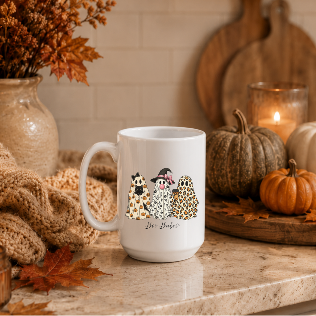 A white ceramic mug with a Halloween-themed graphic of three ghosts in costumes and the text 'Boo Babes' printed on the front, styled on a kitchen counter with autumn decorations including pumpkins, a candle, and a knitted blanket.