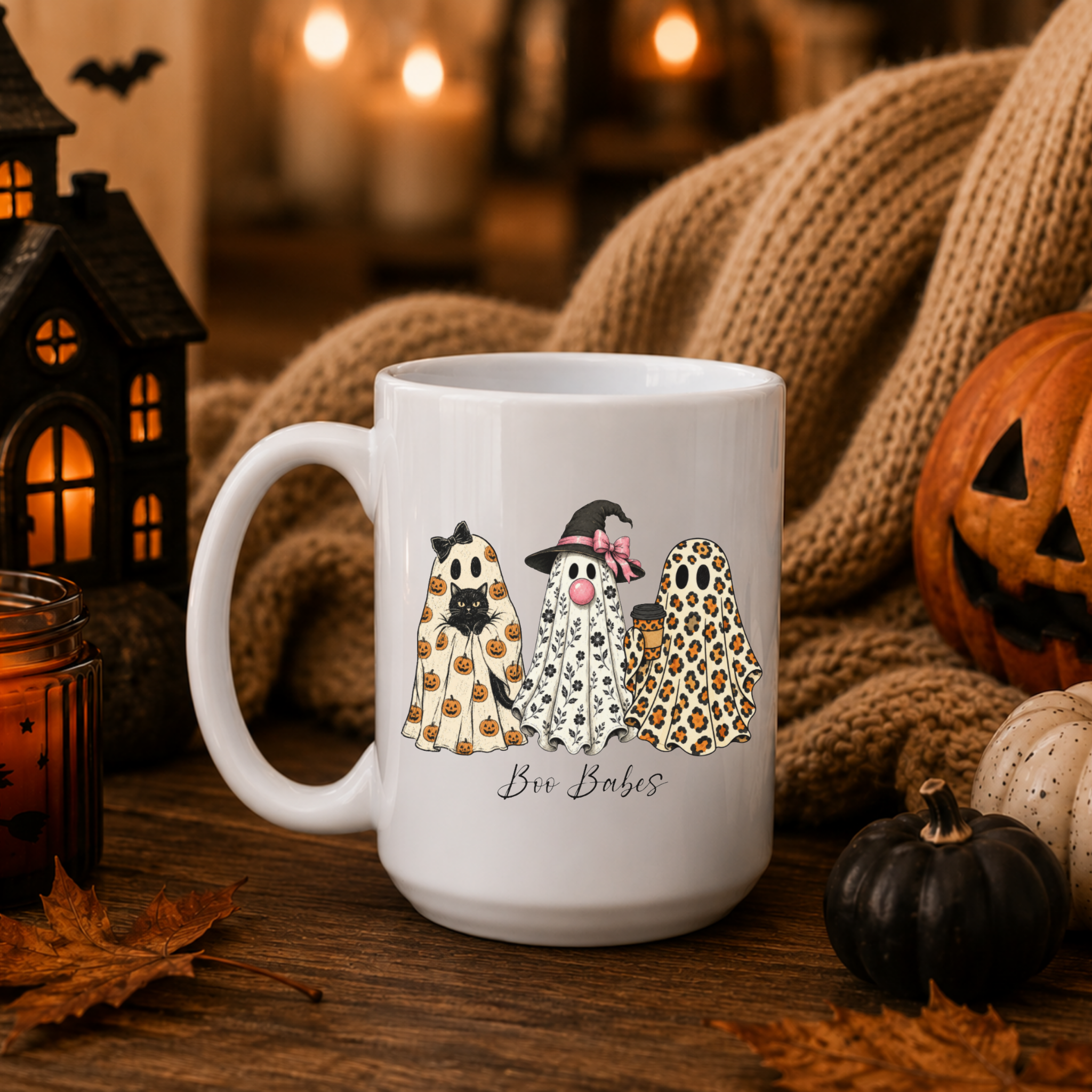 A white ceramic mug with a Halloween-themed ghost illustration and the text 'Boo Babes' printed on it, styled on a wooden table with a knitted blanket, candles, a jack-o'-lantern, and autumn leaves.