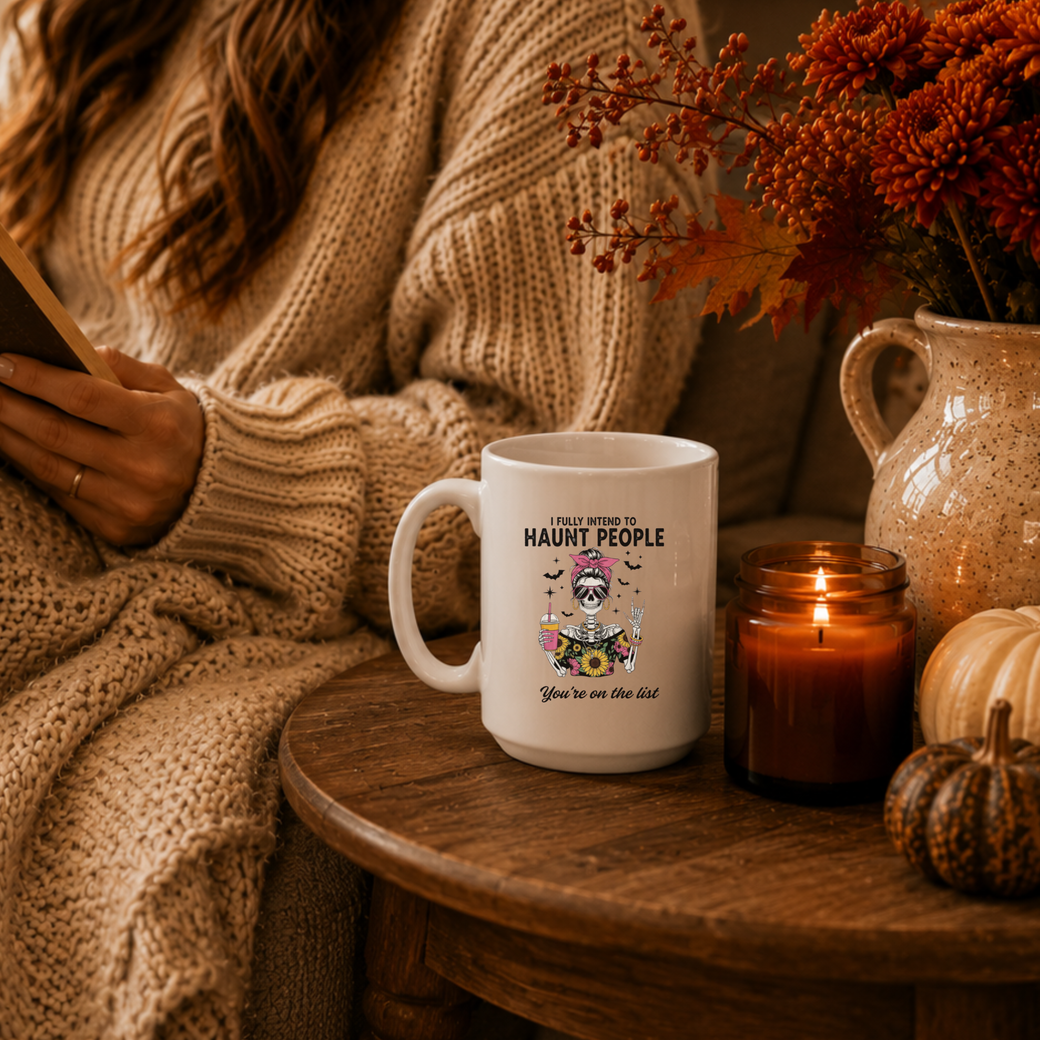 a white ceramic 15 oz mug set on a side table among fall decor. The mug has a fun skeleton with bats and the words "I fully intend to haunt people" and "You're on the list"