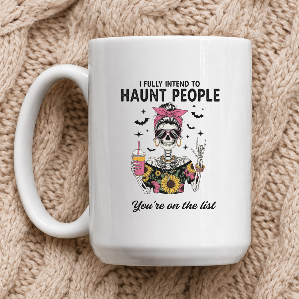 A white ceramic coffee mug with a Halloween graphic of a skeleton woman and the text 'I FULLY INTEND TO HAUNT PEOPLE You're on the list', displayed on a beige knit blanket.
