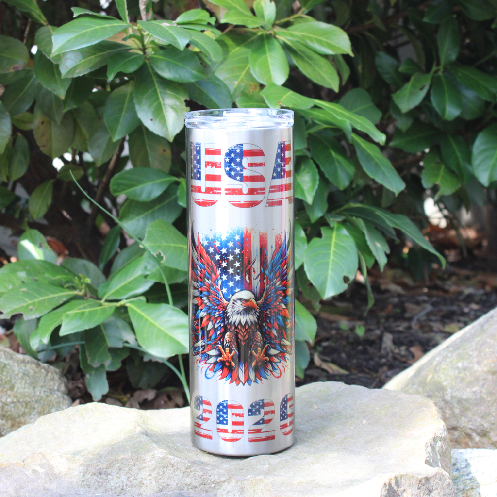 30 oz Stainless Tumbler with USA 2026, American Flag, and Patriotic Eagle on it, sitting on a rock among greenery