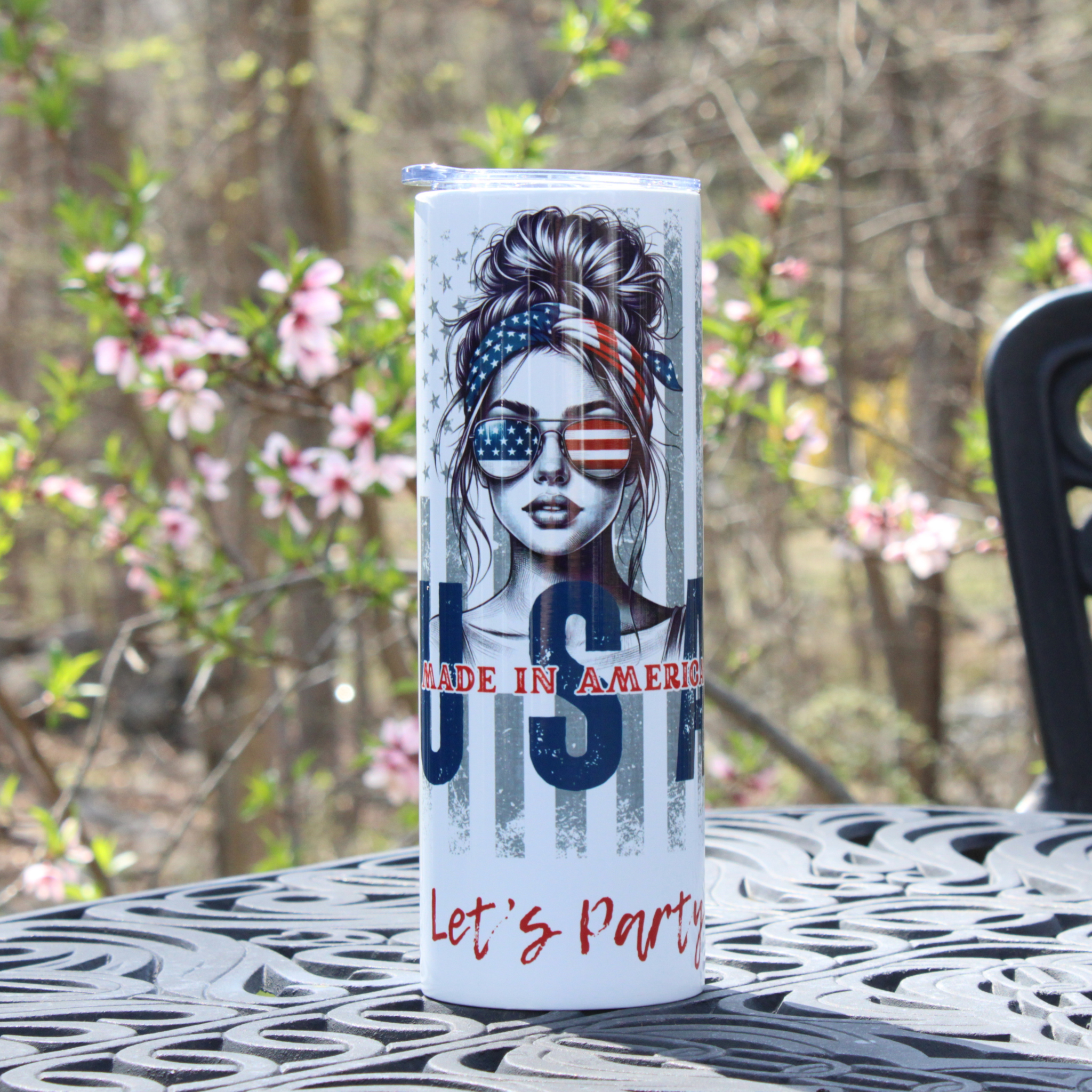 Tumbler with a design of a woman wearing sunglasses and an American flag headband, with 'Made in America' and 'Let's Party' text, set against a blurred natural background.
