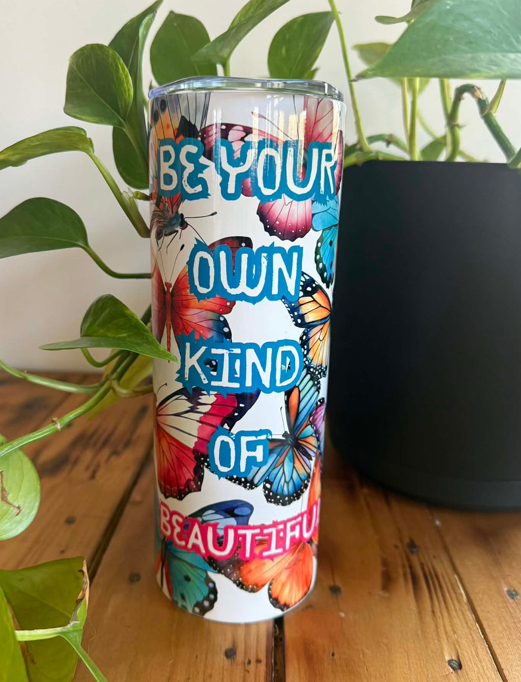 Tumbler with butterfly design and motivational quote on a wooden surface with plants.