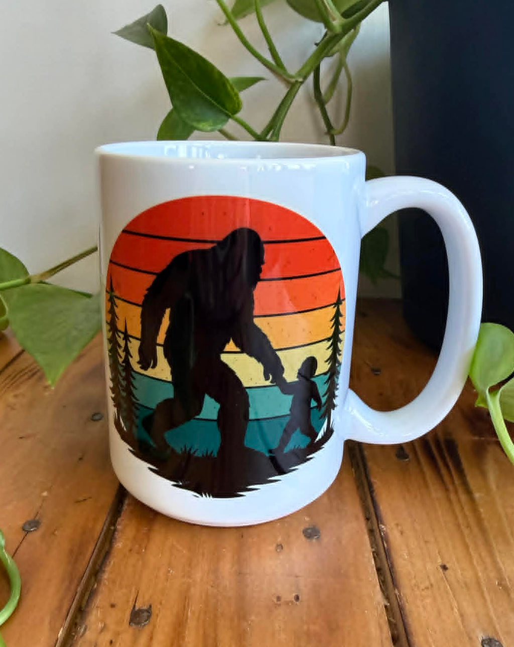 White mug with a Bigfoot design on a wooden surface with plants in the background