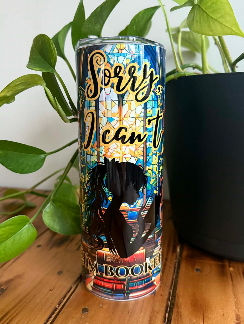 Tumbler with 'Sorry, I can't' text and stained glass design on a wooden surface with a plant in the background